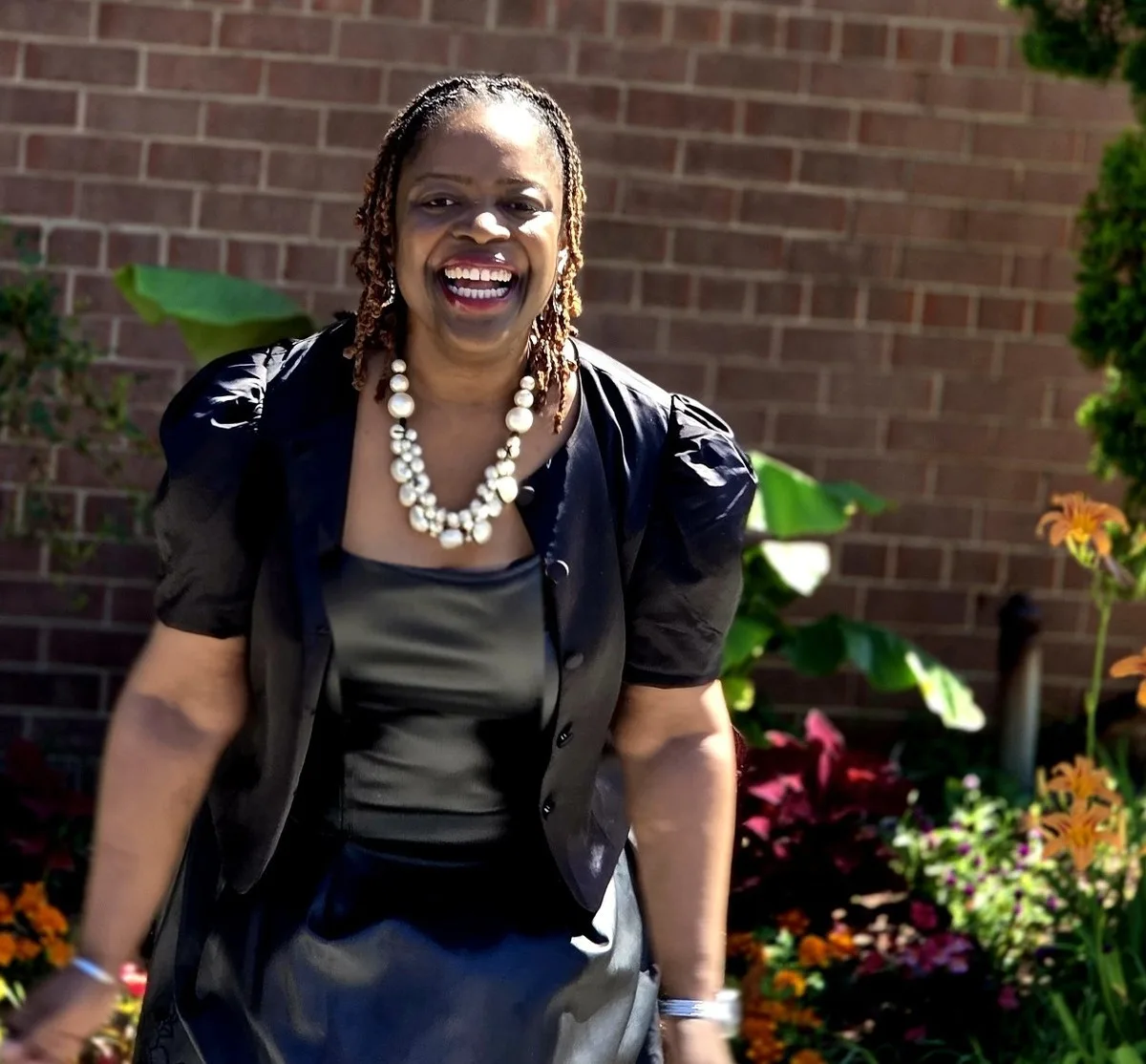 Janelle smiling in a black dress beside a brick wall and flowers
